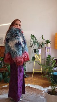 Femme aux longs cheveux portant une veste duveteuse multicolore et une jupe violette, debout dans une pièce avec un sol en bois, des plantes et des tabourets colorés.