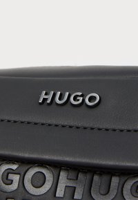 Black leather bag featuring embossed "HUGO" logo and smooth texture, with contrasting stitching and a sleek, rectangular design.