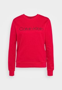 Czerwona bluza wykonana z miękkiej tkaniny, z okrągłym dekoltem, długimi rękawami oraz czarnym logo „Calvin Klein” na przodzie.