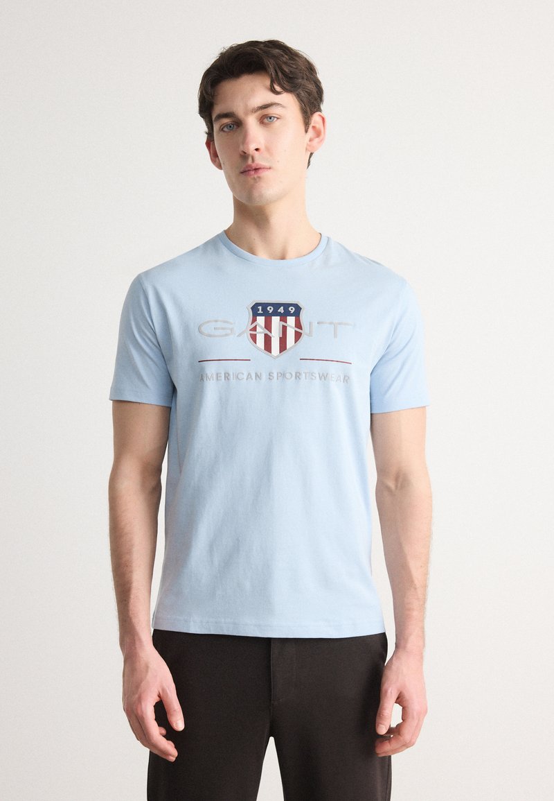 Lyseblå bomulds-T-shirt med et grafisk motiv af et rødt og hvidt skjold, teksten "GANT 1949 AMERICAN SPORTSWEAR" i grå under.
