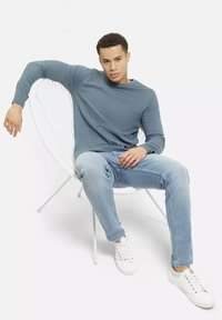 Mężczyzna w niebieskiej koszuli z długim rękawem, jasnych dżinsach i białych trampkach siedzi swobodnie na białym krześle o stalowej konstrukcji na tle jednolitego koloru.