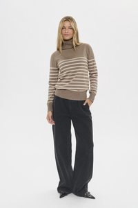 Kobieta stojąca, ubrana w brązowy prążkowany golf, czarne szerokie spodnie i czarne wsuwane buty w szpic z ćwiekami na białym tle.