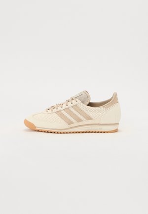 Baskets basses Adidas beige et crème avec trois bandes, empeigne en mesh texturé, semelle en gomme et fermeture à lacets, vue de côté.