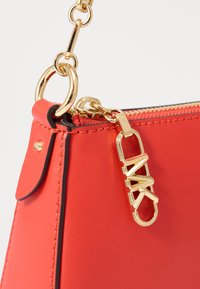 Sac à main en cuir rouge avec des accessoires en or, dotée d'une fermeture éclair et d'un accent de logo décoratif. Texture lisse et forme structurée.