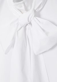 Chemise blanche à col montant avec un grand nœud en tissu noué au cou et des détails de boutons.
