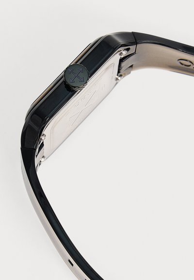 Montre-bracelet rectangulaire noire avec plaque arrière en argent et couronne ronde arborant un logo à quatre flèches sur fond clair.