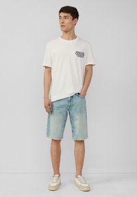 Witte katoenen T-shirt met marineblauw print op de borst, gecombineerd met lichtblauwe denim shorts en off-white sneakers met beige accenten.