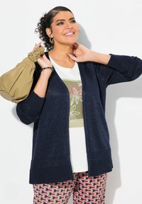 Marineblauer Strickcardigan mit offenem Front, strukturierten Ärmeln, getragen über einem weißen Grafik-T-Shirt. Kombiniert mit gemusterten rosa und schwarzen Hosen.