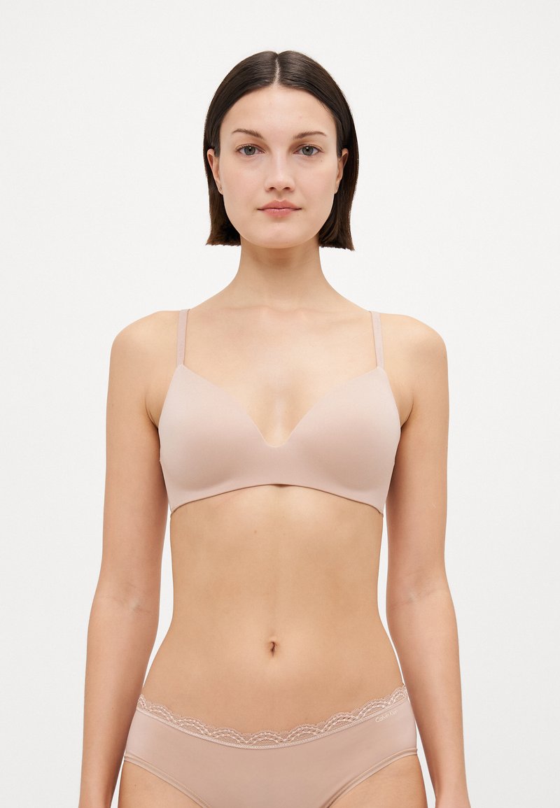 Nude wireless bra with a deep V-neck design, smooth texture, and adjustable thin straps. Notable lace detailing at the waistband of matching briefs.