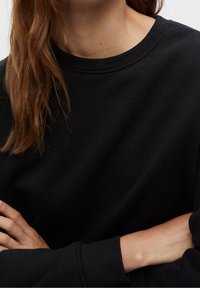 Femme aux longs cheveux bruns portant un pull noir à col rond avec des poignets côtelés, les bras croisés à la taille sur un fond uni.