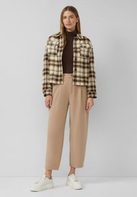 Kariertes Jackett in Creme, Braun und Gelb mit zwei Brusttaschen; kombiniert mit beigen, weit geschnittenen Hosen und weißen Turnschuhen.