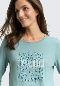 Smiling woman with long brown hair wearing a light blue shirt with a blue patterned square and the word "beautiful" in white letters.