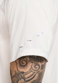 Hellgraues Sportshirt mit glatter Textur, das subtile horizontale Akzente in der Nähe des Ärmels und einem tätowierten Arm im Hintergrund aufweist.