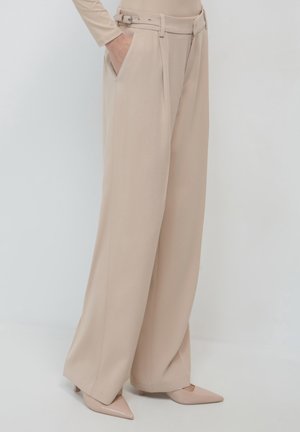 Vrouw draagt beige wijde broek met zijzakken en een dubbele gesp, gecombineerd met beige puntige hakken.