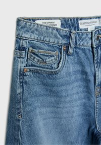 Jean en denim bleu en gros plan montrant la poche avant, petite poche à pièces avec une étiquette métallique, passants de ceinture et étiquettes de marque visibles à l'intérieur de la ceinture.