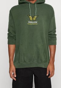 Zelená mikina s kapucí a přední kapsou, s grafikou žlutého motýla a textem "PAPILLONS Botanická výstava." Měkká textilní struktura.
