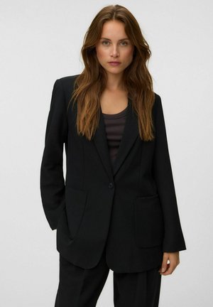Femme aux longs cheveux bruns portant un blazer noir et un pantalon assorti, debout avec une main dans la poche devant un fond uni.