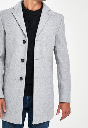 Homme portant un manteau en laine gris clair boutonné par-dessus un pull noir et un jean bleu, debout devant un fond blanc uni.