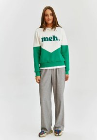Zelená a bílá mikina s textem "meh." v kombinaci se šedými širokými kalhotami a vícebarevnými botami na světlém pozadí.