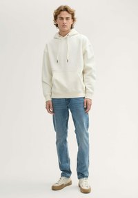 Sudadera blanca con capucha y cordones, bolsillo frontal y sutil detalle de logo. Combinada con jeans azul claro y zapatillas beige.