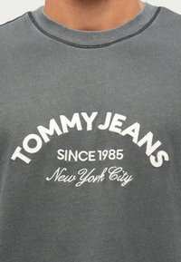 Γκρι βαμβακερό t-shirt με λαιμόκοψη και κεντημένο κείμενο: "TOMMY JEANS" που ανεβαίνει πάνω από "SINCE 1985" και "New York City" από κάτω.