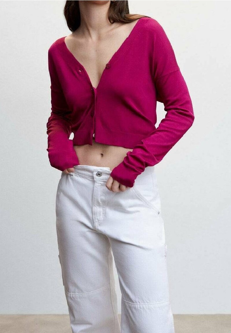 Femme portant un cardigan boutonnière magenta court et un pantalon blanc taille haute, les mains près du waistband.