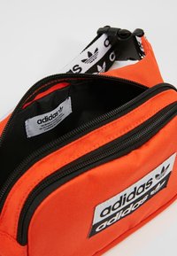 Orange fabric bag with black accents, featuring a zippered opening, dual compartments, and branded labels inside and on the front.