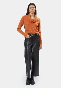 Oranje gebreide trui met een col, gecombineerd met zwarte leren wijde broek. De outfit wordt aangevuld met een zwarte riem met metalen oogjes.