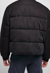 Chaqueta tipo puffer negra con costuras horizontales, un cuello alto y puños elásticos. El material parece suave y acolchado, proporcionando aislamiento.