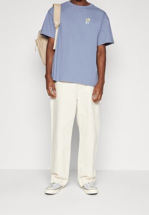 T-shirt in cotone azzurro chiaro con una piccola grafica floreale, abbinato a pantaloni a gamba larga color crema e sneakers grigie. È indossato uno zaino beige.