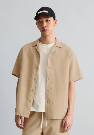 Junger Mann trägt ein beiges Kurzarmhemd mit Knöpfen über einem weißen T-Shirt, beige Hose, silberne Kette und schwarze Kappe vor grauem Hintergrund.