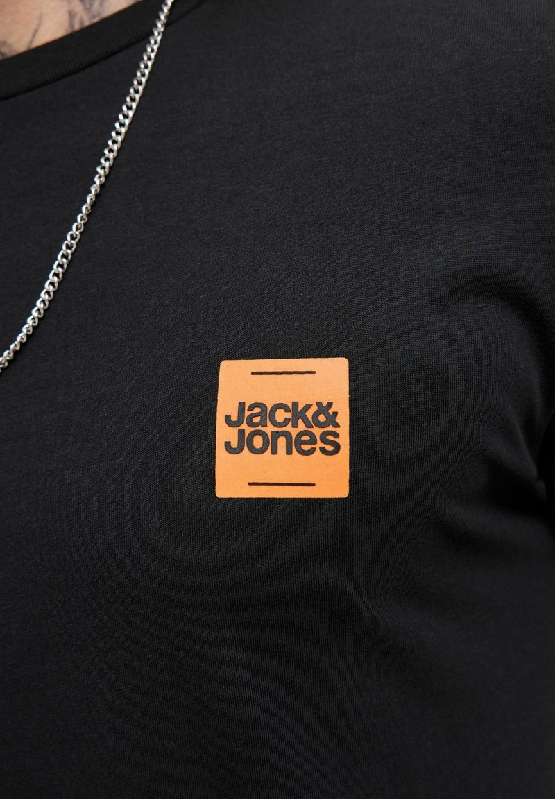 Černé tričko s oranžovým čtvercovým logem "Jack & Jones" na hrudi, doplněné stříbrným náhrdelníkem.