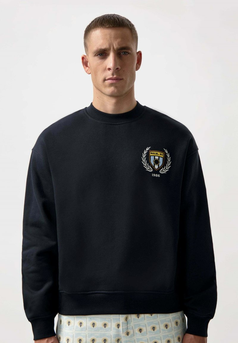 Granatowy sweter crewneck z haftowanym emblematem featuring wieniec laurowy i tekst "BALR. FC 1986." Miękka tkanina, luźny krój, minimalistyczny design.