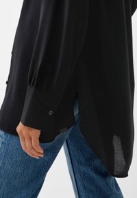 Camicia nera a maniche lunghe con tessuto testurizzato, polsini con bottoni e spacchi laterali, abbinata a jeans in denim azzurro chiaro.