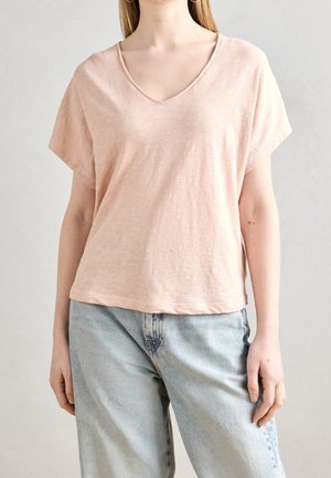 Frau trägt ein blassrosa V-Ausschnitt Kurzarmshirt und hellblaue hochtaillierte Jeans, steht vor einem einfarbigen hellen Hintergrund.