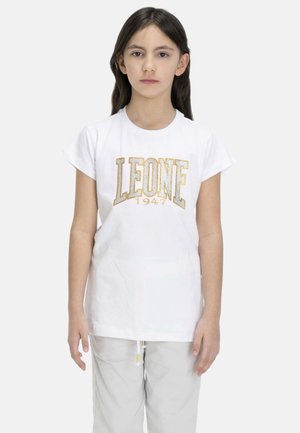 Une fille se tient debout, face à l’avant, sur un fond uni. Elle porte un tee-shirt blanc avec l’inscription dorée « LEONE 1947 » et un pantalon clair.