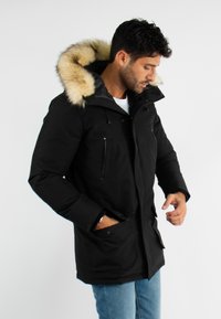 Parka noire avec capuche en fausse fourrure, poches zippées, cordons de serrage ajustables et textures variées. Le mannequin porte un jean bleu clair.