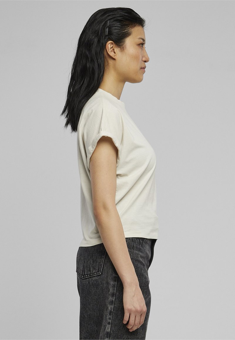 Young woman with long black hair wearing a cream short-sleeve shirt and dark gray jeans, standing in profile against a gray background.