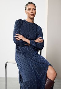 Marineblaules, gepunktetes Kleid mit langen Ärmeln und einem kleinen Schlitz. Glatte Textur, lockere Passform. Model sitzt auf einem Chromstuhl.