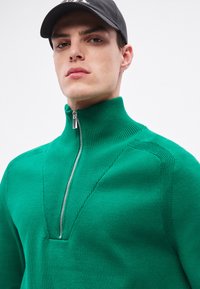 Zielony sweter na zamek z prążkowanym kołnierzem i teksturowanymi detalami na ramionach. Gładka dzianina o dopasowanym kroju z srebrnym zamkiem błyskawicznym.