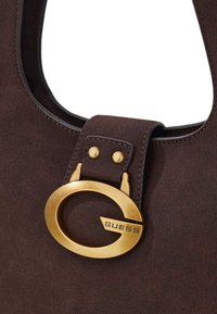 Borsa a tracolla in camoscio marrone con un emblema circolare dorato e dettagli metallici. Presenta un bordo cucito e una tracolla rigida strutturata.