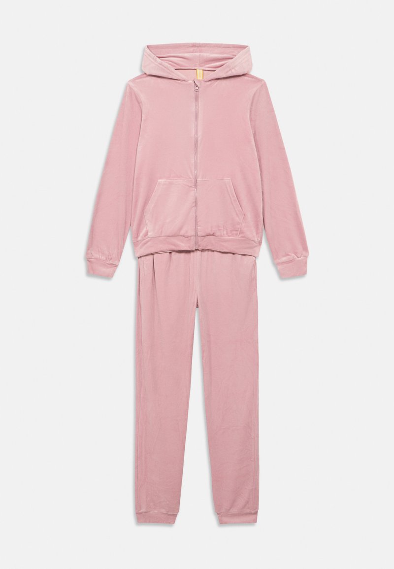 Rožinės spalvos veliūrinis sportinis kostiumas, kuriame yra gobtuvas su užtrauktuku ir priekine kišene bei derančios kelnės su riboto audinio apvadais. Visas kostiumas turi minkštą tekstūrą.