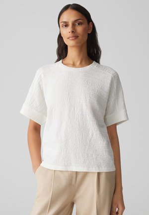 Witte gestructureerde t-shirt met korte mouwen en een ronde halslijn. De stof heeft een gekreukt patroon en is gecombineerd met beige broeken.