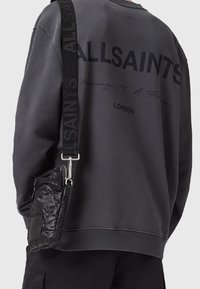 Personne portant un sweat-shirt ALLSAINTS gris foncé avec un sac à bandoulière noir avec une sangle portant la marque ALLSAINTS, vue de dos.