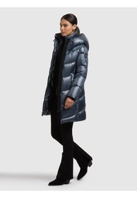 Ilgas, blizgus pūkinis paltas tamsiai mėlynos spalvos, su gobtuvu, užtrauktuku priekyje ir su siuvine dekoru. Dėvimas ant juodo marškinėlio ir kelnių.