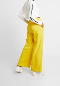 Lösa gula byxor med sidofickor, i kombination med en vit långärmad topp med svarta grafiska detaljer på ärmarna.