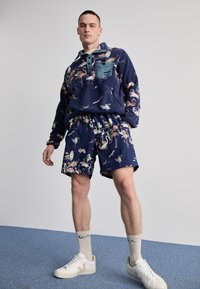 Marineblå genser med vid bunn, mønstret med flamingoer, sammen med matchende shorts. Hvite sneakers med beige detaljer og stripete sokker.