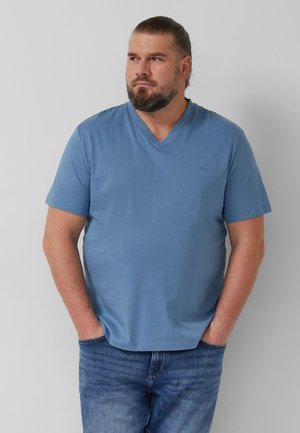 Brodaty mężczyzna z długimi włosami w niebieskiej, krótkawej koszulce z dekoltem w serek i niebieskich dżinsach, stojący z rękami w kieszeniach i spoglądający w lewo.