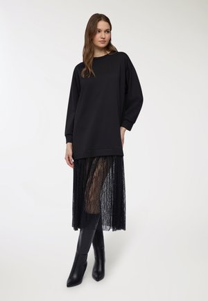 Felpa nera oversize con maniche lunghe, indossata sopra una gonna nera in pizzo trasparente. Stivali neri alti fino al ginocchio con punta appuntita completano l'outfit.