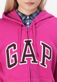 Fukcija dukserica na raskopčavanje u boji fuksije s natpisom "GAP" izvezena u bijeloj i tamnosmeđoj boji. Nosena preko plave i zelene karirane košulje s dugmetom na ovratniku.